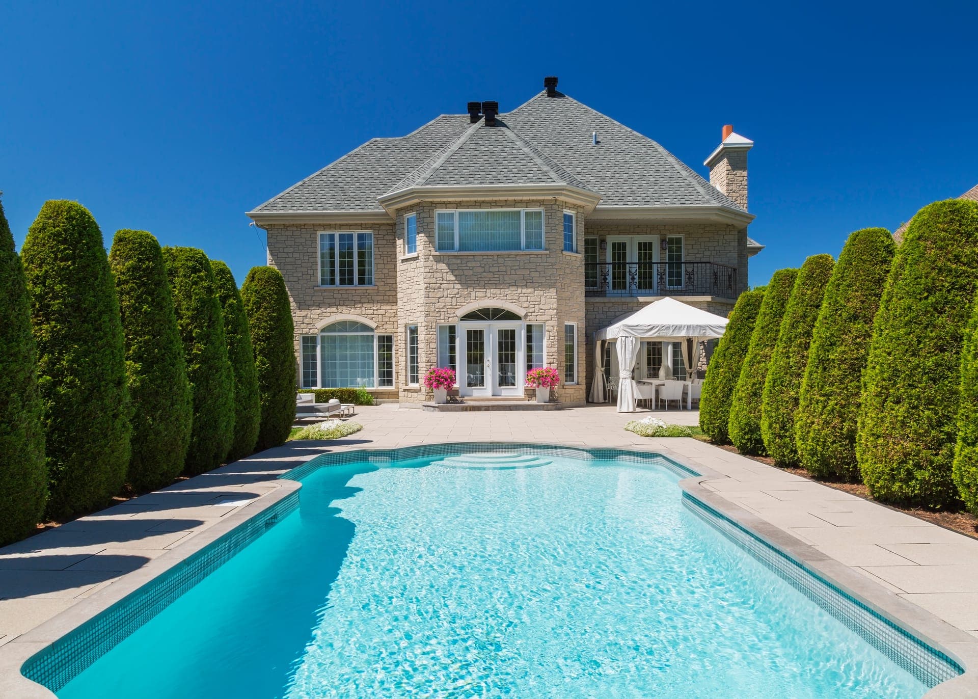 Large stone home with an in-ground pool, paved terrace and trimmed cedar hedges