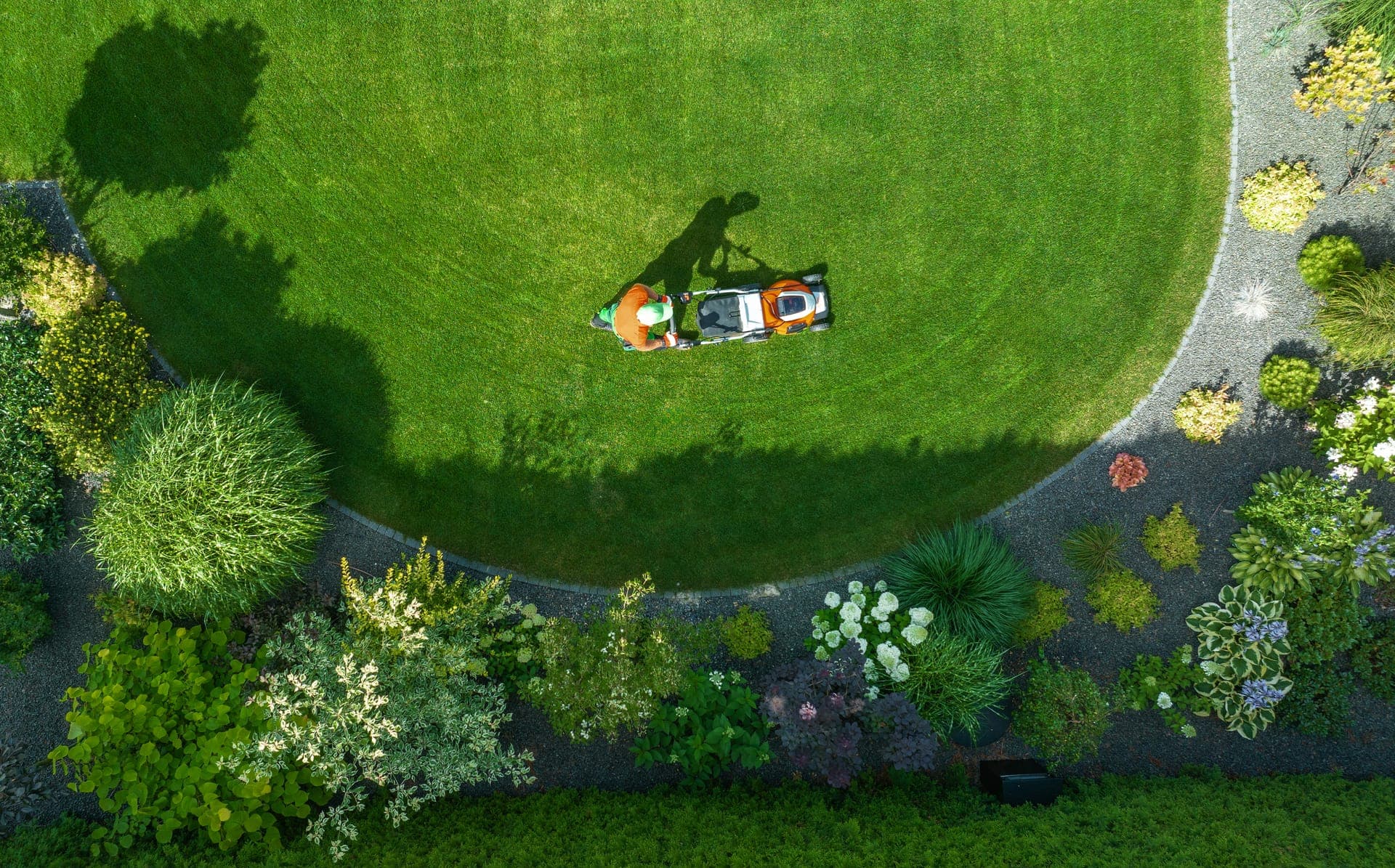 Aerial view of a person mowing a large curved lawn edged with planted beds and a gravel path