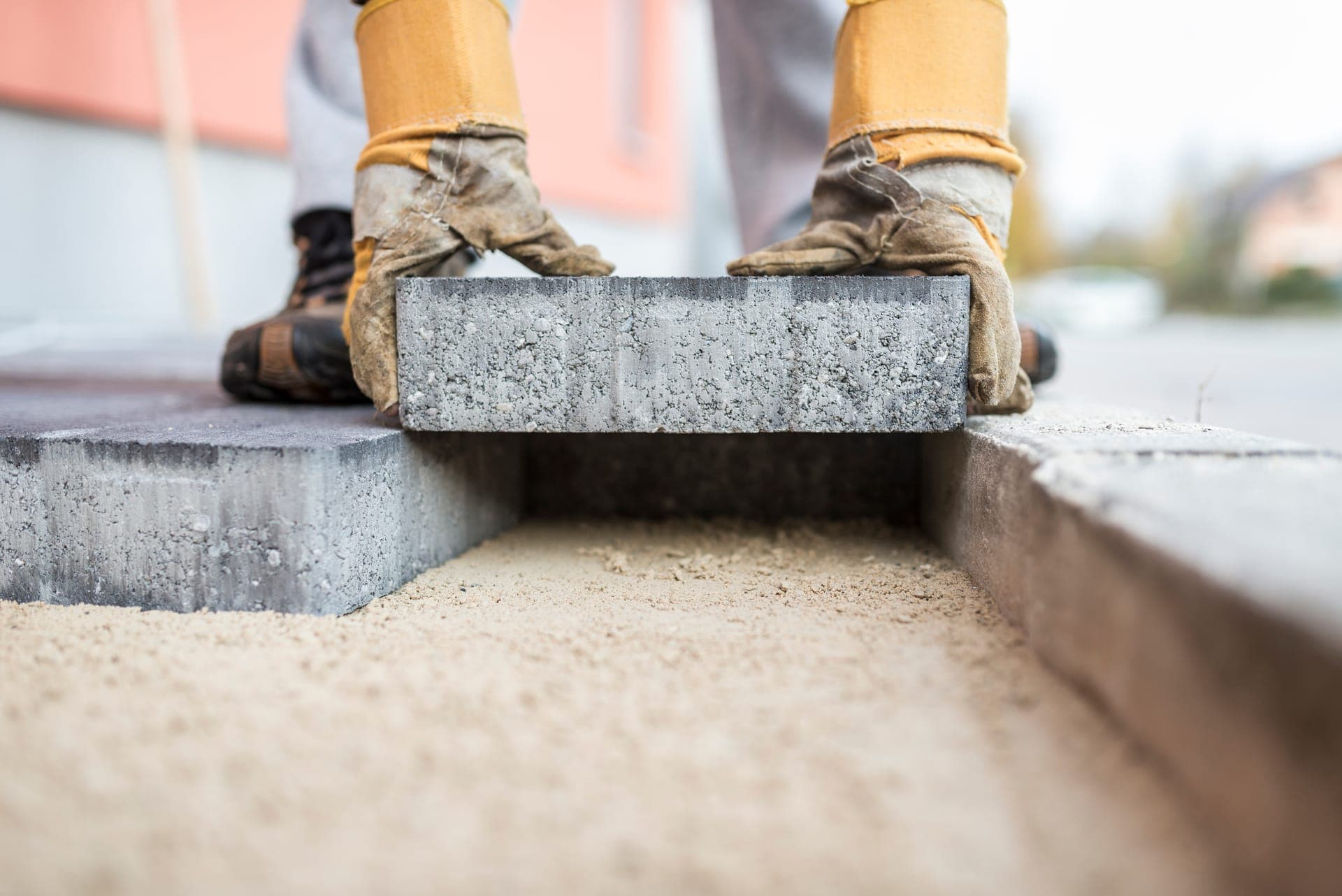 Setting a grey concrete paver onto a bed of levelled sand, gloved hands